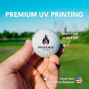 a hand holding a golf ball with a logo on it, set against a backdrop of a grassy field and a clear blue sky.