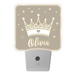 May include: A beige night light with a white crown and stars design. The name "Olivia" is written below the crown. The night light has a white plug-in base with an on/off switch.