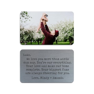 May include: A silver metal card with a photo of a man holding a child in the air. The card has a message that reads "Luke, We love you more than words can say. You're our everything. Your love and make our home complete. Your biggest fans are always cheering for you. Love, Mindy + Amanda"
