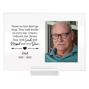 May include: A white memorial plaque with a black border and a photo of a man with glasses. The text reads: "Those we love don't go away. They walk beside us every day. Unseen, Unheard, But Always Near. Still Loved, Still Missed and very Dear. Dad 1955 - 2023"