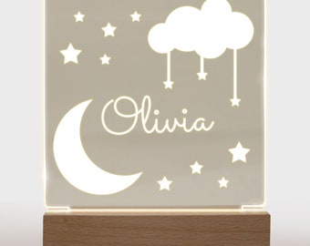 May include: A white acrylic night light with a crescent moon, a cloud with stars hanging from it, and the name "Olivia" in a cursive font. The light is on a wooden base.
