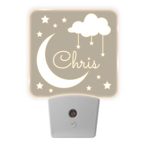 May include: A white night light with a beige plastic cover. The cover has a white crescent moon, a cloud with stars hanging from it, and the name "Chris" written in a cursive font. The night light is illuminated, casting a warm glow.