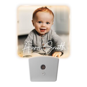 May include: A night light featuring a smiling baby with red hair and the name "Aaron Smith" in white script. The night light has a square, glowing face and a white base with an on/off button.