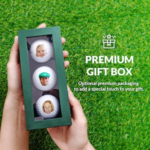 A person's hands holding a green gift box containing three golf balls with faces on them, set against a grassy background.