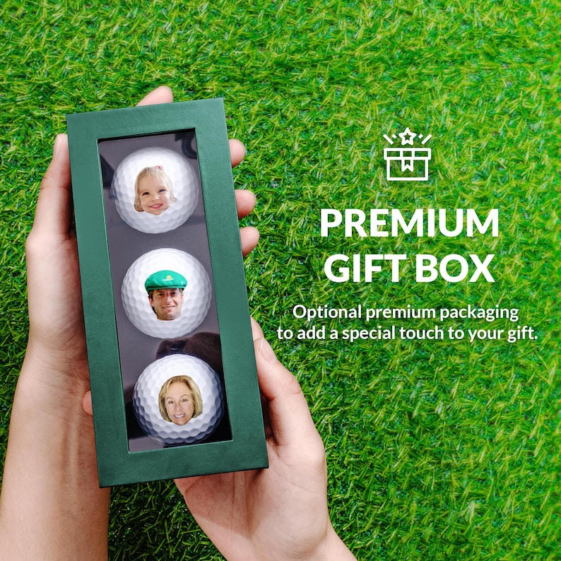 A person's hands holding a green gift box containing three golf balls with faces on them, set against a grassy background.