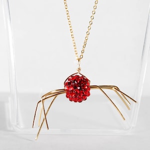 May include: A gold chain necklace with a red jeweled spider pendant. The spider has eight legs and a round body.