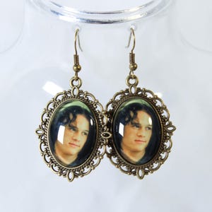Op de afbeelding: Een paar bronzen oorhangers. Elke oorbel heeft een ovaal portret in een decoratief filigraan frame. De portretten tonen een persoon met donker haar. De oorbellen hangen aan een eenvoudige haak.