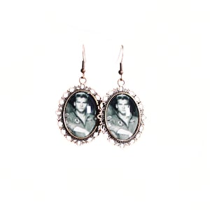May include: A pair of vintage-style earrings. Each earring features an oval-shaped, black and white image of a man in a military uniform, set within an ornate, bronze-colored frame. The earrings hang from simple fish hook ear wires.