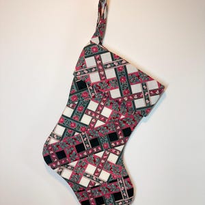 May include: A holiday stocking featuring a woven-look pattern of intersecting bands in cream, black, and dark green. The design includes floral accents in shades of pink and green throughout the fabric, finished with a matching fabric hanging loop.