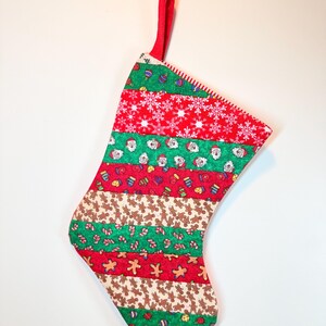 May include: A Christmas stocking constructed from horizontal fabric strips featuring festive patterns including white snowflakes on red, Santa Claus faces on green, gingerbread men, candy canes, and colorful ornaments on various backgrounds.