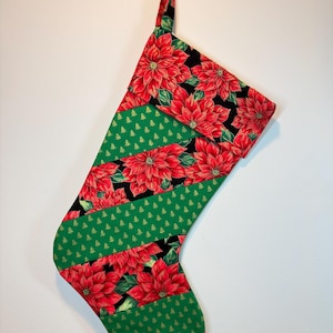 May include: A Christmas stocking featuring a diagonal patchwork design. The fabric alternates between a black background with red poinsettia flowers and green leaves, and a solid green fabric patterned with small, repeating gold Christmas trees.