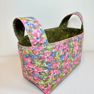May include: A rectangular fabric basket with two arched handles. The exterior features a floral pattern with pink, blue, and white flowers on a white background. The interior and handles are a solid green color.