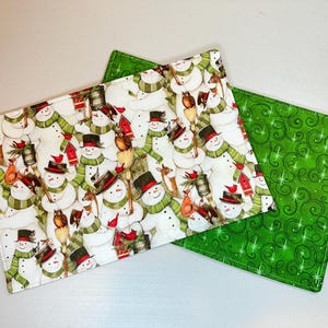 May include: Two rectangular Christmas-themed placemats. One features a pattern of snowmen with top hats, scarves, and birds. The other is solid green with a swirling pattern. The placemats are likely made of fabric.