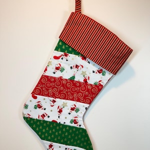 May include: A Christmas stocking featuring a red and white striped cuff and hanging loop. The body consists of alternating horizontal bands of white fabric with a Santa Claus and star print, a red fabric with gold swirls, and green with trees.