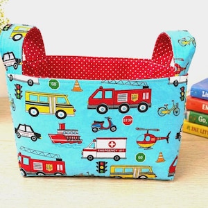 May include: A rectangular fabric storage basket with handles. The exterior is light blue with a pattern of emergency vehicles, traffic lights, and bicycles. The interior is red with white polka dots. A stack of children's books is visible.