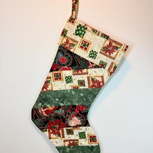 May include: A fabric Christmas stocking featuring a patchwork design with alternating diagonal stripes. The pattern includes cream, black, and green sections decorated with red poinsettias, green stars, and floral motifs. It has a hanging loop at the top.