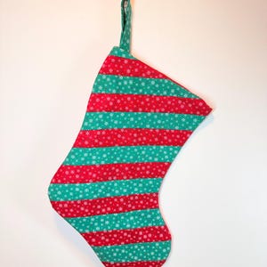 May include: A fabric Christmas stocking featuring alternating horizontal stripes of red and green. Both the red and green fabric sections are patterned with small, white, six-pointed snowflake designs. A matching fabric loop is attached at the top.