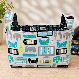 May include: A rectangular fabric storage basket with two handles. The basket is patterned with video game controllers, screens, and the text "NEXT LEVEL OVER" in teal, gray, and black on a white background. The handles have a striped design.