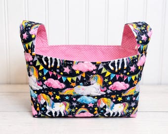 Peut inclure: Un panier de rangement en tissu avec un extérieur noir orné d'un motif de licorne coloré, une doublure rose à pois et deux poignées. Les licornes ont des crinières et des queues arc-en-ciel, avec des nuages, des étoiles et des arcs-en-ciel.