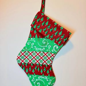 May include: A fabric Christmas stocking featuring a patchwork design of alternating horizontal bands. The pattern includes red fabric with green pine trees, bright green fabric with white swirling flourishes, and a red, green, and white plaid.