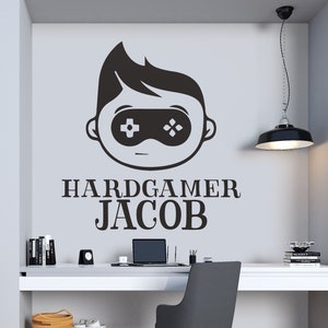 K&ouml;nnte beinhalten: Ein modernes Heimb&uuml;ro mit einem Wandtattoo mit einem Gamer-Motiv und dem Text "HARDGAMER JACOB". Der Raum umfasst einen wei&szlig;en Schreibtisch, einen grauen Stuhl und Topfpflanzen. Der Boden ist aus Holz.