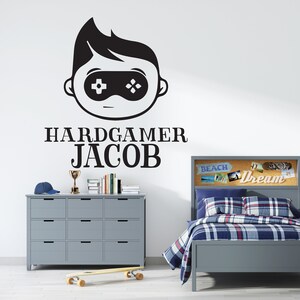 K&ouml;nnte beinhalten: Graues Schlafzimmer-Set mit Bett, Kommode und Nachttisch. Das Bett hat eine blau-rot karierte Bettdecke. Ein Wandtattoo zeigt eine Gamer-Grafik und den Namen "Jacob". Ein Skateboard und eine Troph&auml;e sind ebenfalls zu sehen.
