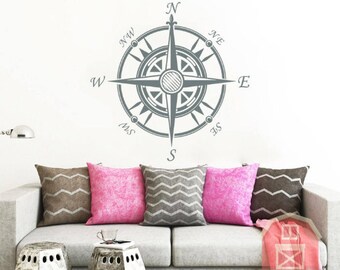 May include: A gray compass wall decal with the points of the compass labeled N, NW, W, SW, S, SE, E, and NE. The compass is on a white wall above a gray couch with pillows.