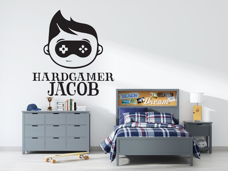 K&ouml;nnte beinhalten: Graues Schlafzimmer-Set mit Bett, Kommode und Nachttisch. Das Bett hat eine blau-rot karierte Bettdecke. Ein Wandtattoo zeigt eine Gamer-Grafik und den Namen "Jacob". Ein Skateboard und eine Troph&auml;e sind ebenfalls zu sehen.