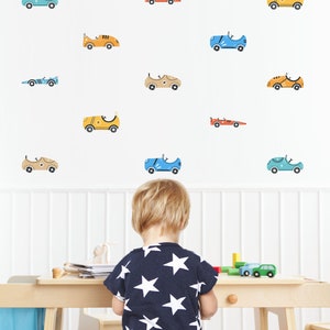 May include: A young child stands in front of a white wall with colorful car decals. The child is wearing a navy blue shirt with white stars and is looking at a table with wooden toys.