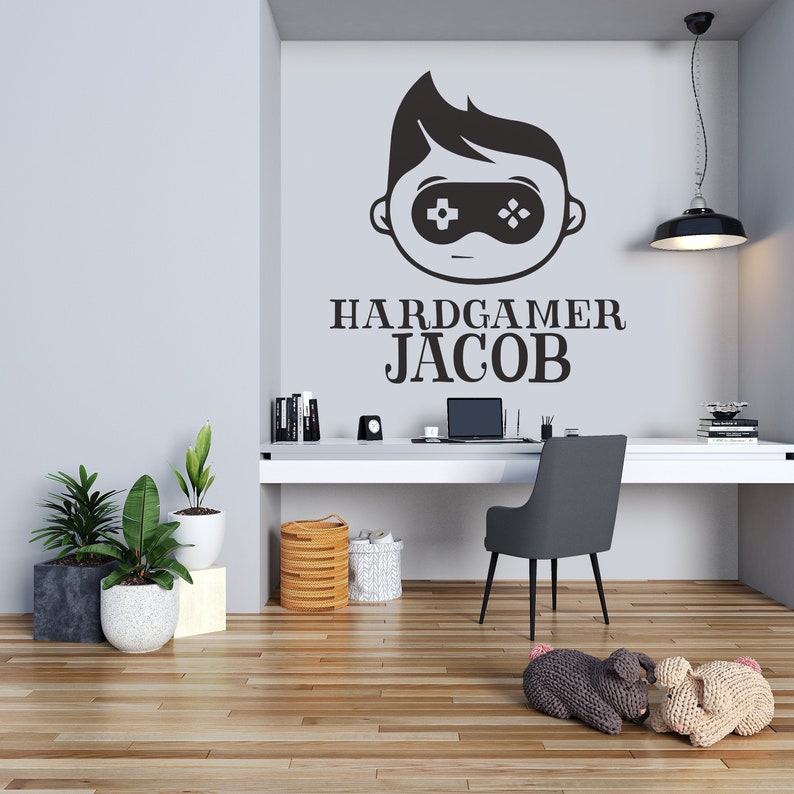 K&ouml;nnte beinhalten: Ein modernes Heimb&uuml;ro mit einem Wandtattoo mit einem Gamer-Motiv und dem Text "HARDGAMER JACOB". Der Raum umfasst einen wei&szlig;en Schreibtisch, einen grauen Stuhl und Topfpflanzen. Der Boden ist aus Holz.