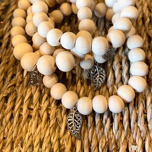 May include: Three bracelets made of light-coloured wooden beads, each with a silver leaf charm. The round beads are strung together. The leaf charms are intricately designed. The bracelets are presented on a woven, textured surface.