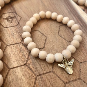 May include: A wooden bead bracelet with a gold bee charm. The bracelet is made of natural wood beads and has a simple, elegant design. The bee charm is adorned with sparkling rhinestones.
