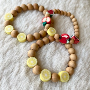 May include: Two wooden bracelets with fruit charms. One bracelet has lemon charms and the other has apple, watermelon, and cherry charms.
