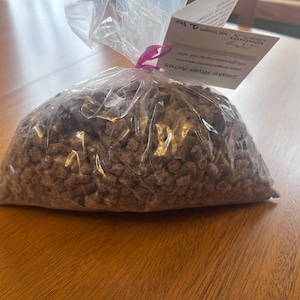 May include: A clear plastic bag filled with brown, granular material. A tag attached to the bag reads "Snake River Acres" and "Net Weight 2 lb". The bag is tied with a pink ribbon and sits on a wooden surface.
