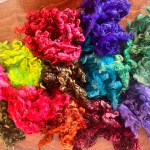 May include: A collection of colorful, curly wool roving in shades of pink, red, orange, yellow, green, blue, and purple. The wool is arranged in fluffy, textured clumps, ideal for fiber art projects like spinning, felting, or weaving.