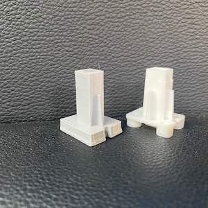 May include: Two white plastic 3D printed objects. The object on the left is taller and has a rectangular base. The object on the right is shorter and has a square base with four legs.