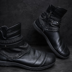 Könnte beinhalten: Ein Paar schwarze Lederstiefeletten mit seitlichem Reißverschluss, einem umlaufenden Riemen mit Metallniete und einem strukturierten Schaft. Die Stiefel haben eine abgerundete Zehenpartie, eine flache Sohle und sichtbare Nähte am Obermaterial.