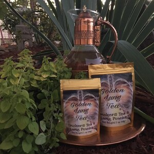 Pu&ograve; includere: Due bustine dorate di Golden Lung Tea, una miscela di t&egrave; per fumatori e polvere per i polmoni. Le bustine sono su una superficie color rame con vegetazione verde sullo sfondo. Il testo sulle bustine recita "Golden Lung Tea" e "Smokers Tea & Lung Powder". La bustina pi&ugrave; piccola &egrave; etichettata "Peso netto 1 oz (28 g)" e la bustina pi&ugrave; grande &egrave; etichettata "Peso netto 2 oz (56 g)".