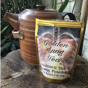 Pu&ograve; includere: Una bustina dorata con l'immagine di un polmone sul davanti. La bustina contiene Golden Lung Tea & Lung Powder. La bustina &egrave; appoggiata su una superficie di legno accanto a una teiera marrone.