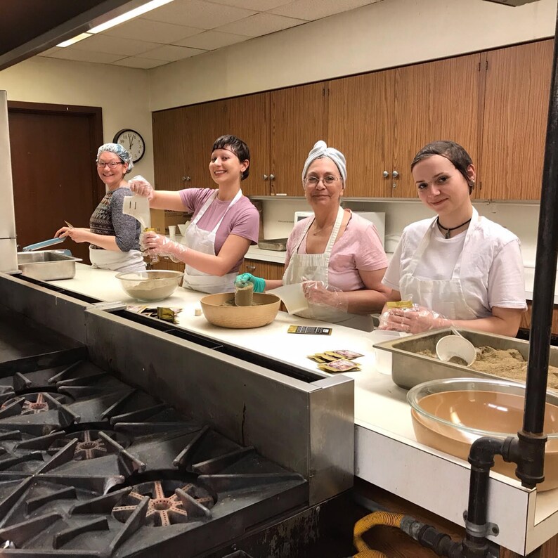 Pu&ograve; includere: Quattro persone con grembiuli bianchi e guanti stanno preparando cibo in una cucina. Sono in piedi a un bancone con un lavello in acciaio inossidabile e un fornello a gas. Ci sono ciotole di ingredienti sul bancone, tra cui una ciotola di polvere marrone.