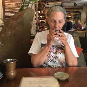 Pu&ograve; includere: Una donna indossa una maglietta bianca con il testo "olden Lung Tea" e un'immagine di una tigre. Sta tenendo una piccola tazza nera e sorseggiando del t&egrave;. Il tavolo &egrave; color rame e c'&egrave; un piccolo piattino bianco con un disegno floreale.