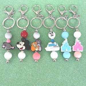 Könnte beinhalten: Eine Sammlung von sechs Schlüsselanhängern mit silberfarbenen Beschlägen. Jeder Schlüsselanhänger hat einen anderen Anhänger, darunter Zeichentrickfiguren und ein Kreuzfahrtschiff, mit bunten Perlen. Die Anhänger sind an einem Ring und einem Verschluss befestigt.