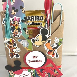 May include: A brown paper envelope-style holder decorated with Disney character stickers including Mickey Mouse, Minnie Mouse, and Goofy. It contains a bookmark with a light blue tassel, a red pen, a pencil, and a bag of Haribo Goldbears.
