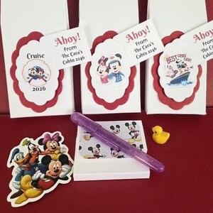 Può includere: Tre sacchetti regalo rettangolari bianchi con etichette smerlate rosse con grafiche di personaggi Disney e testo tra cui Ahoy! From The Cara's Cabin 2246. Include una penna glitter viola, una piccola papera di gomma gialla e un adesivo Disney.