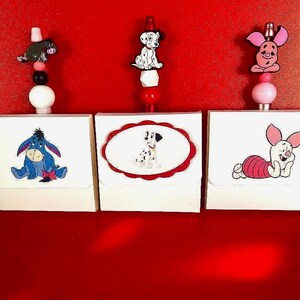 May include: Three white rectangular boxes featuring cartoon character illustrations of Eeyore, a Dalmatian puppy, and Piglet. Each box is topped with a vertical beaded topper featuring a matching character charm in black, white, red, and pink colors.