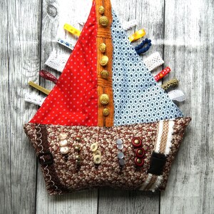 Puede incluir: Un juguete sensorial de tela hecho a mano con forma de barco, con una vela roja de lunares, una vela azul estampada y una base floral marrón. Cuenta con botones, cintas y varias texturas para la exploración táctil. Botones dorados en el centro. Para el desarrollo infantil temprano.