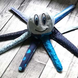 Puede incluir: Un juguete de peluche con forma de pulpo con una cara sonriente y ojos azules. El pulpo tiene ocho tentáculos estampados en tonos azules, incluyendo lunares, flores y diseños de olas. El juguete está hecho de tela suave.