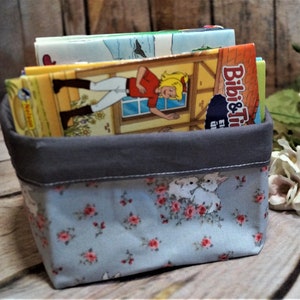 Peut inclure: Un panier de rangement en tissu gris et bleu avec un motif floral et des lapins blancs. Le panier est rempli de plusieurs livres pour enfants colorés, dont un avec le titre " Bibi & Tina".