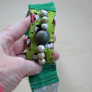 Puede incluir: Una diadema de tela verde y blanca con una fila de cuentas plateadas y verdes y una cuenta verde grande en el centro. La tela tiene un patrón de mariquitas rojas sobre un fondo verde.