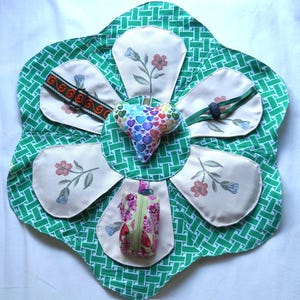 May include: A handmade, flower-shaped organizer with a green and white patterned background. It features six cream-colored petal-shaped pockets, each embroidered with a floral design. A heart-shaped pillow with colorful hearts is in the center, and a small zippered pouch is attached.